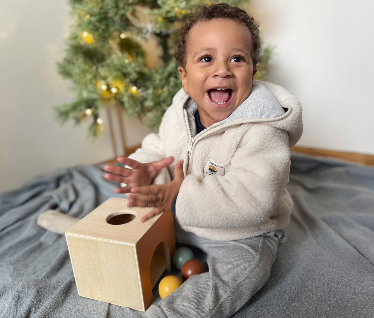 Ein glückliches Kleinkind in einer cremefarbenen flauschigen Jacke sitzt auf einer grauen Decke, lachend mit erhobenen Händen, neben einem small foot Montessori-Bundle 3 Spielzeug aus Holz mit bunten Bällen und einem verschwommenen Weihnachtsbaum im Hintergrund.