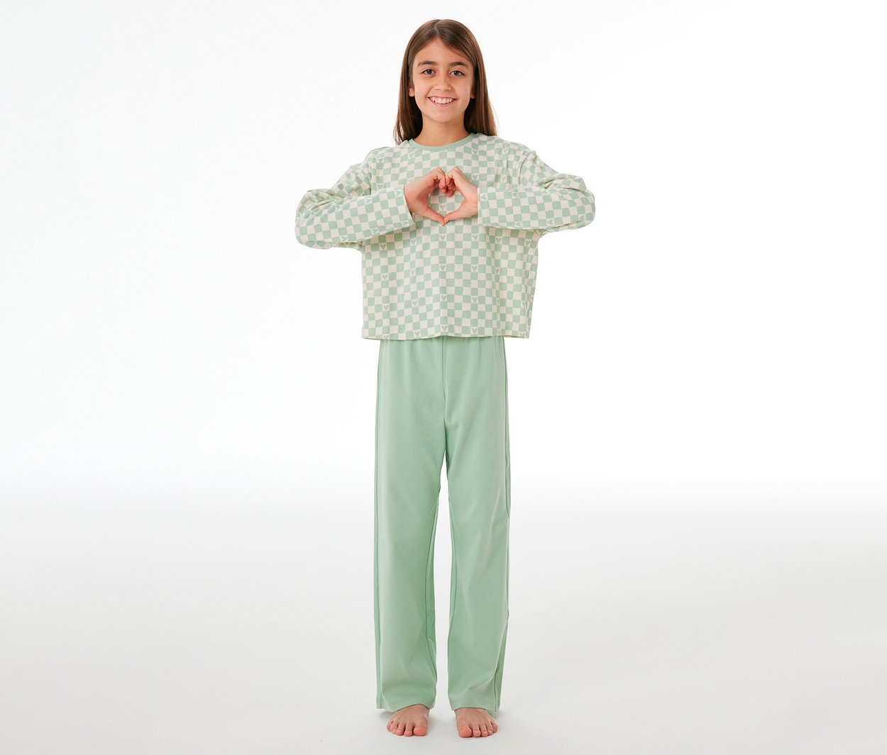 Lächelndes junges Mädchen in einem SCHIESSER Kinder-Interlock-Pyjama, das mit den Händen ein Herz formt. Sie trägt ein hellgrün-weiß kariertes Langarmshirt und eine einfarbige hellgrüne lange Hose.