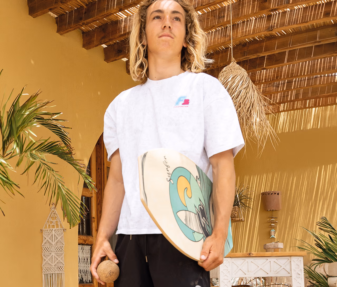 Junger Mann mit blonden Locken hält ein F2 Balance Board Drop mit Wellenmotiv und einen Ball, stehend vor einer gelben Wand mit Palmen.
