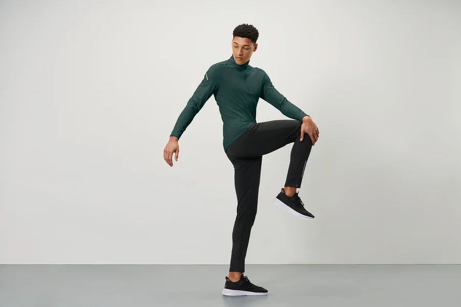 Mann in grünem Sportoberteil und schwarzen Windprotection-Lauftight hält ein Knie mit einer Hand vor der Brust.