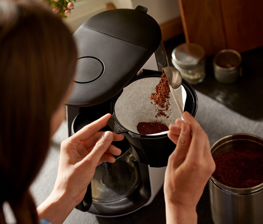 Frau füllt mit einem Löffel Kaffee in den weißen Filter der »Let's Brew« Filterkaffeemaschine.