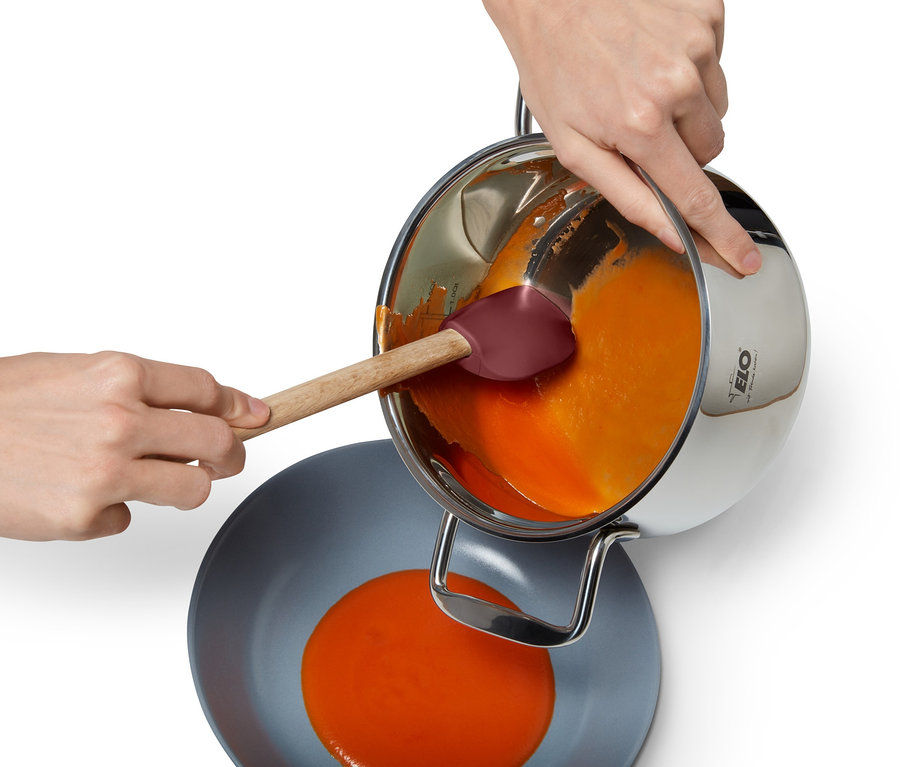Eine Person gießt eine orangefarbene Flüssigkeit aus einem Edelstahltopf in eine graue Schüssel und verwendet dabei einen Multifunktions-Kochlöffel.