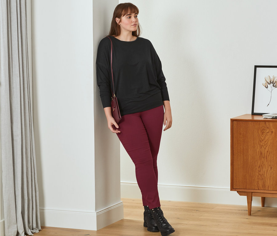 Eine Frau lehnt an einer Wand und trägt ein schwarzes Langarmshirt mit Satindetail am Ausschnitt, eine dunkelrote Stretchhose, Leder-Boots und eine rote Mini-Tasche.