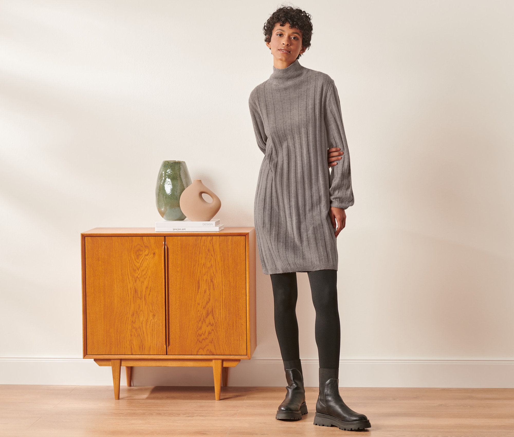 Frau posiert neben einem Schrank und trägt ein geripptes Strickkleid mit Stehkragen, eine schwarze Leggings und Stiefel.