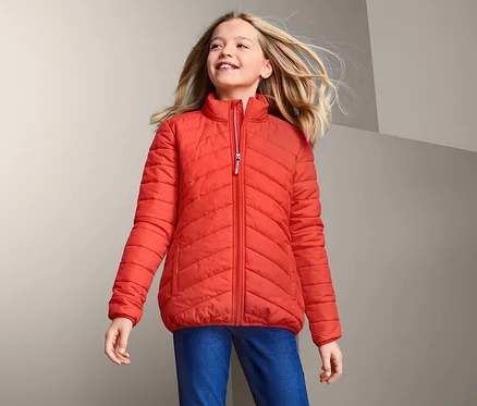 Lächelndes Mädchen trägt eine orangefarbene Steppjacke mit recyceltem Material und schaut nach oben.