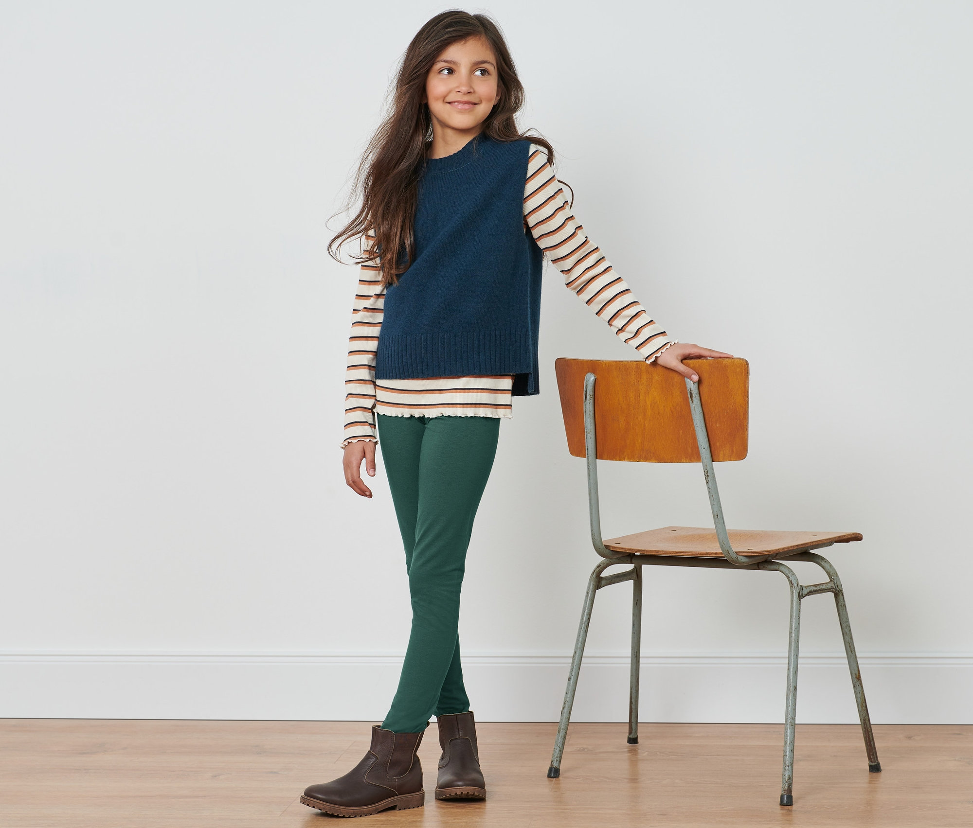 Mädchen steht und lehnt an einem Stuhl, trägt grüne 3 Leggings und braune Stiefel.