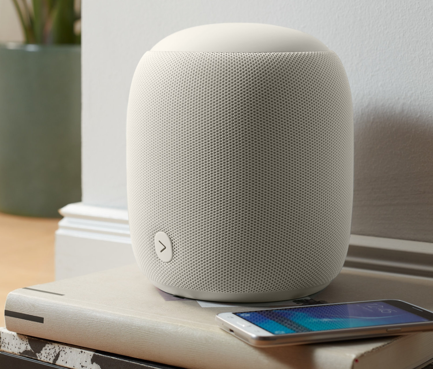 Ein grauer Fabric-Bluetooth®-Lautsprecher steht auf einem Stapel Bücher neben einem Mobiltelefon.