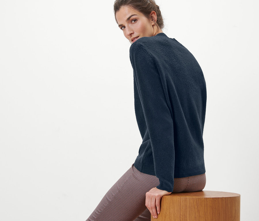 Frau sitzt auf einem Holzhocker und trägt einen Strickpullover in Boucléoptik.