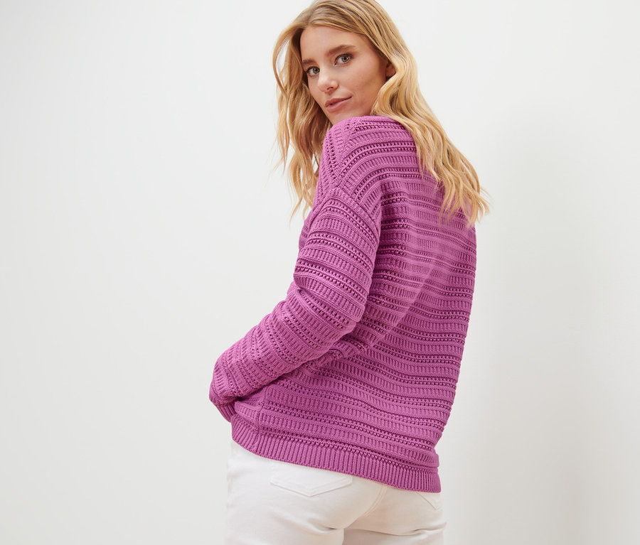 Blonde Frau blickt über die Schulter und posiert in einem Pullover mit Ajour-Muster und einer weißen Hose.