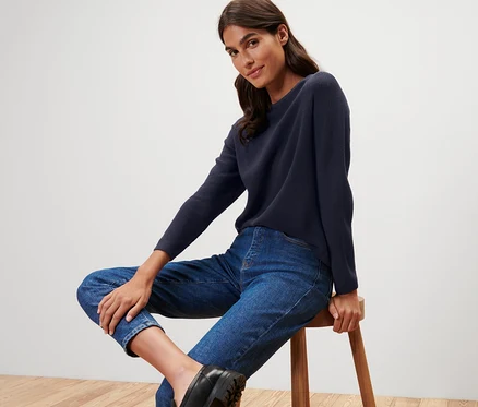 Eine Frau sitzt auf einem Hocker und trägt einen dunkelblauen Strickpullover.