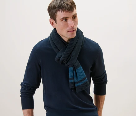 Mann mit Strickschal, marineblauem Pullover und Jeans, der sich an einen Stuhl lehnt.