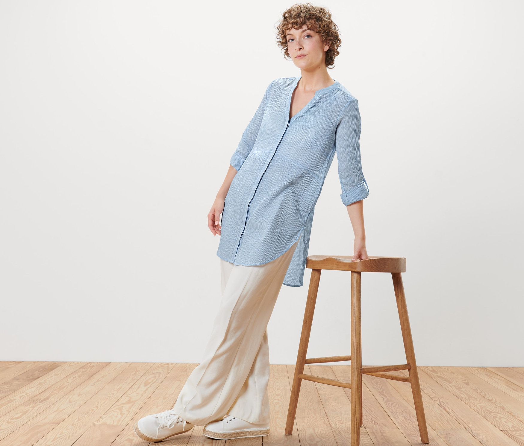 Eine Frau, die sich an einen Holzhocker lehnt, trägt eine gestreifte Longbluse.