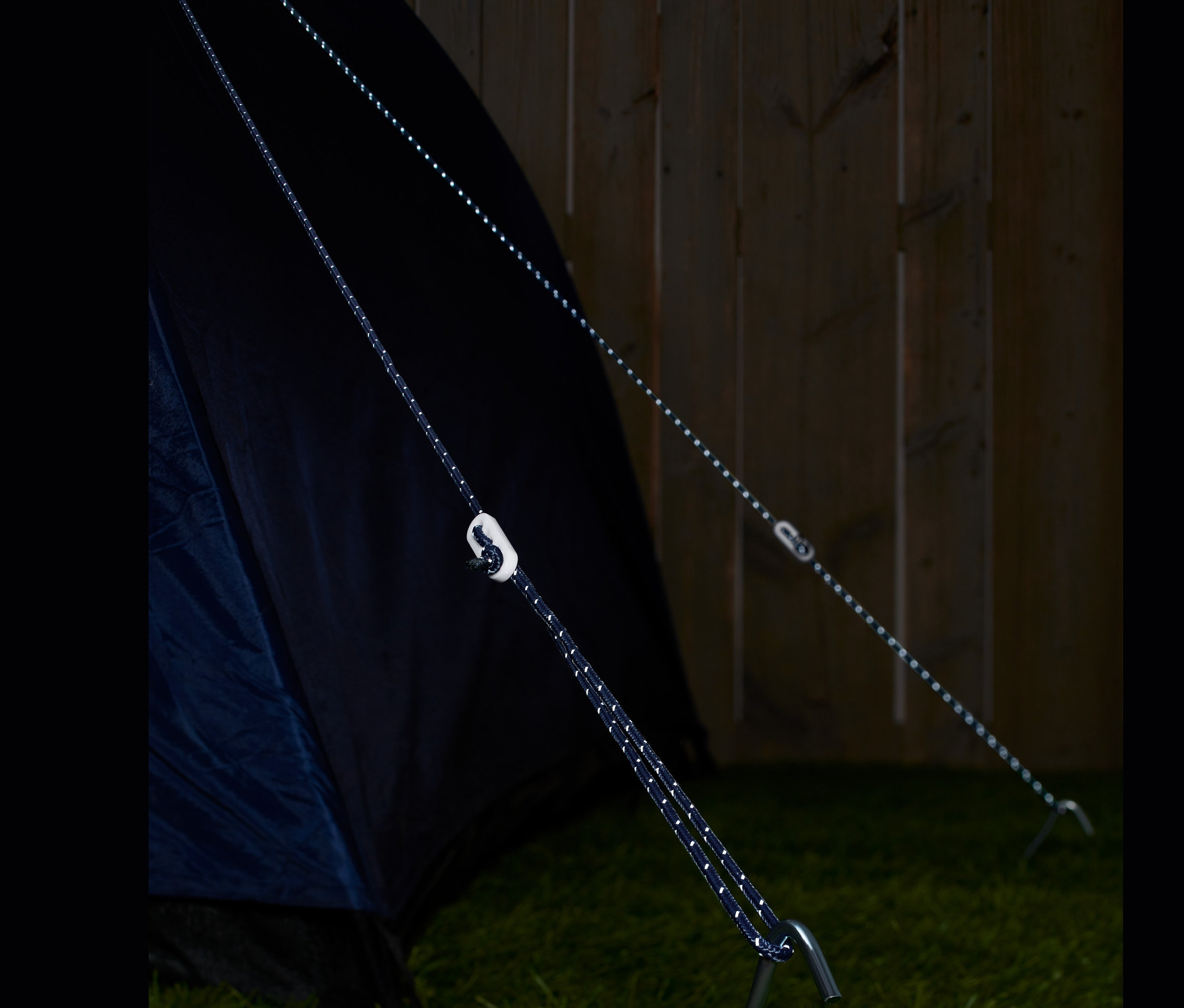 Zwei reflektierende Abspannseile sind an einem Zelt und einem Hering im Gras befestigt.