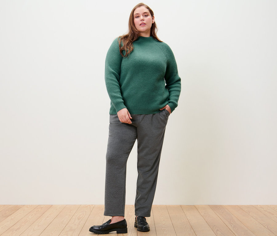 Frau posiert in grünem Grobstrickpullover mit Stehkragen und Joggpants.