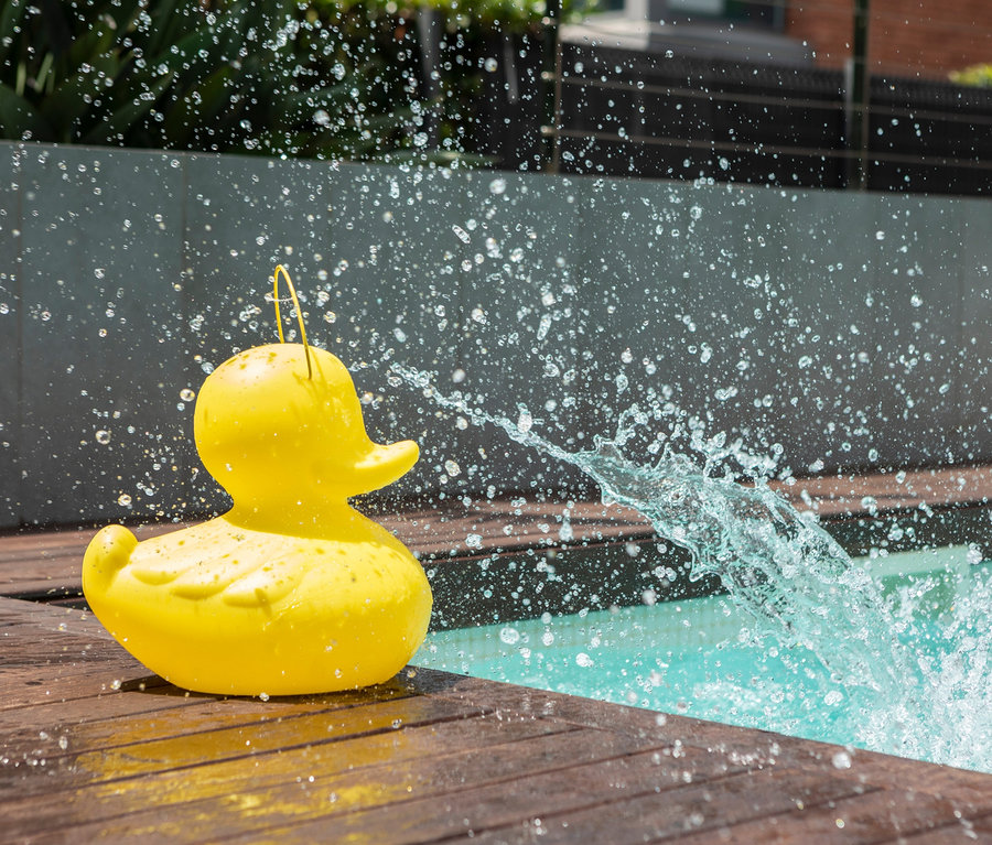 Eine gelbe Goodnight Light LED-Leuchte »Ente« S steht auf einem Holzboden neben einem Pool, der mit Wasser bespritzt wird.