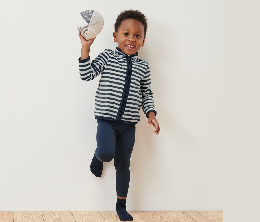 Ein Junge hält einen Stoffball hoch und trägt eine Kinder-Fleecejacke mit Ringeln.