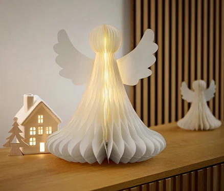 Ein großer LED-Papier-Engel steht auf einer Holzkommode neben einem beleuchteten Keramikhaus.