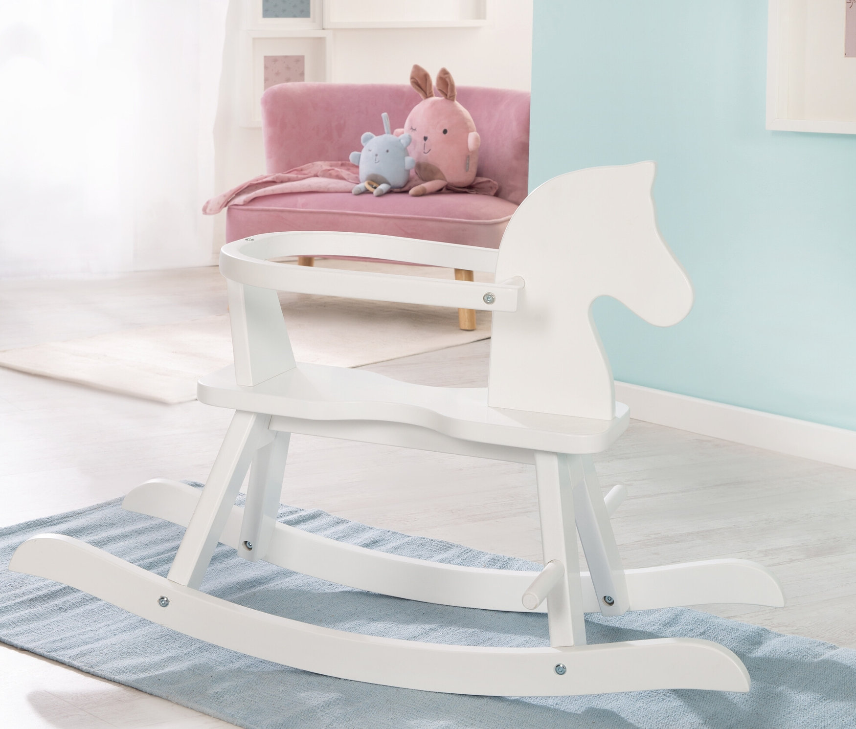 Ein weißes Schaukelpferd steht auf einem blauen Teppich. Im Hintergrund befinden sich ein rosa Sofa und ein Plüschtier. roba Schaukelpferd