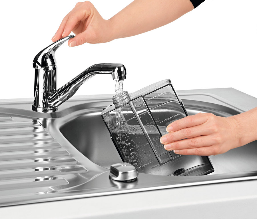 Eine Frau befüllt einen durchsichtigen Behälter mit Wasser aus einem Wasserhahn in einem Edelstahlspülbecken.