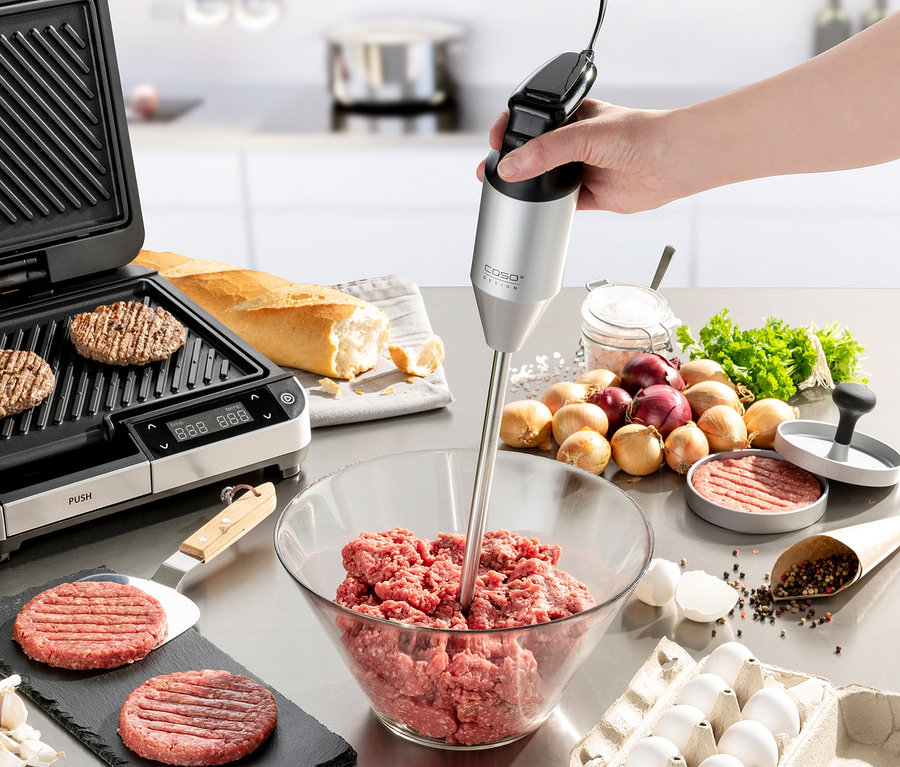 Eine Hand benutzt einen CASO Stabmixer »HB 2400 Pro XL« in einer Schüssel mit Hackfleisch. Auf dem Tisch liegen ein Grill, ein Brötchen, Zwiebeln und Eier.