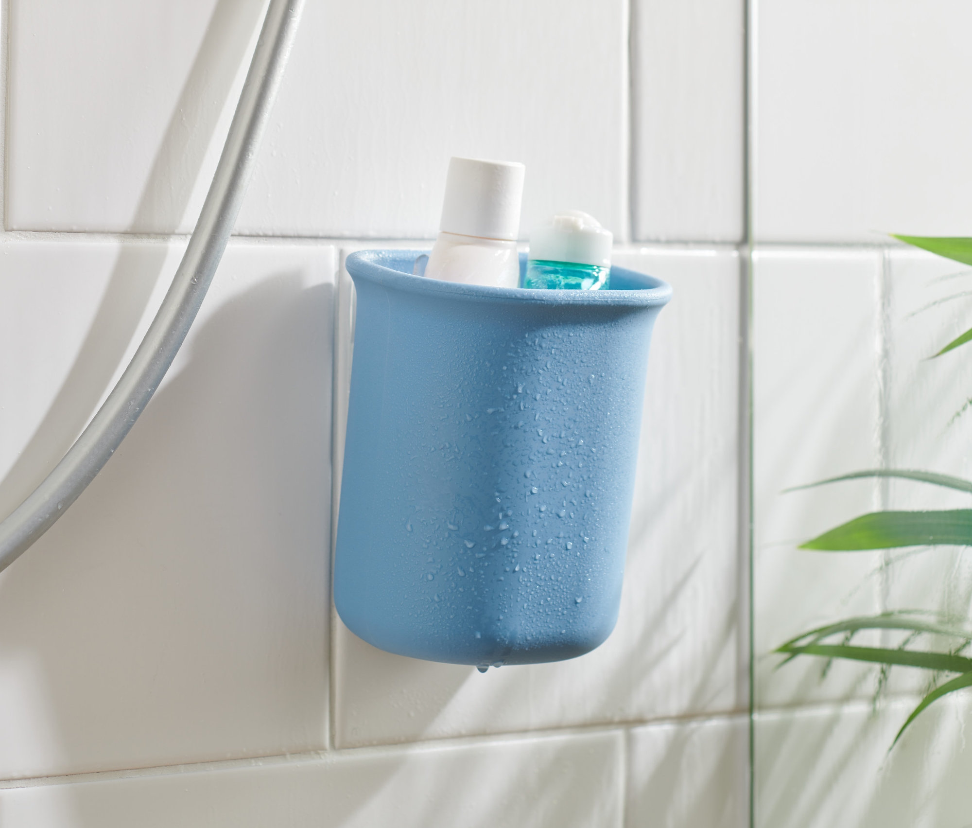 Ein blauer Bad-Organizer mit einem weißen und einem blauen Produkt darin, an weißen Fliesen neben einem Duschschlauch befestigt.