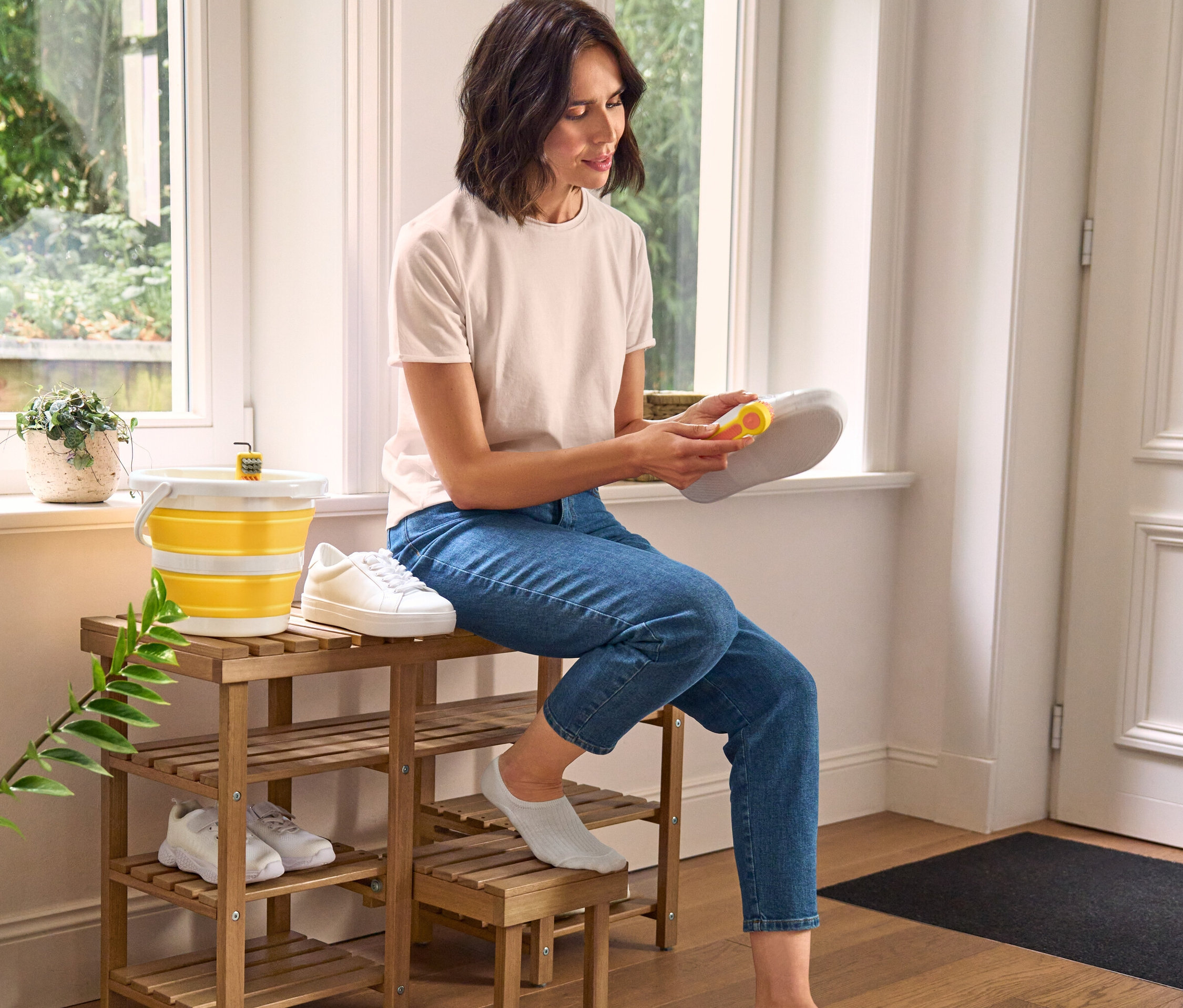 Frau putzt Schuh mit 3-in-1-Schuhreinigungsbürste auf hoher Schuh-Sitzbank mit Klapptritt. Daneben stehen Schuhbürste und faltbarer Putzeimer.