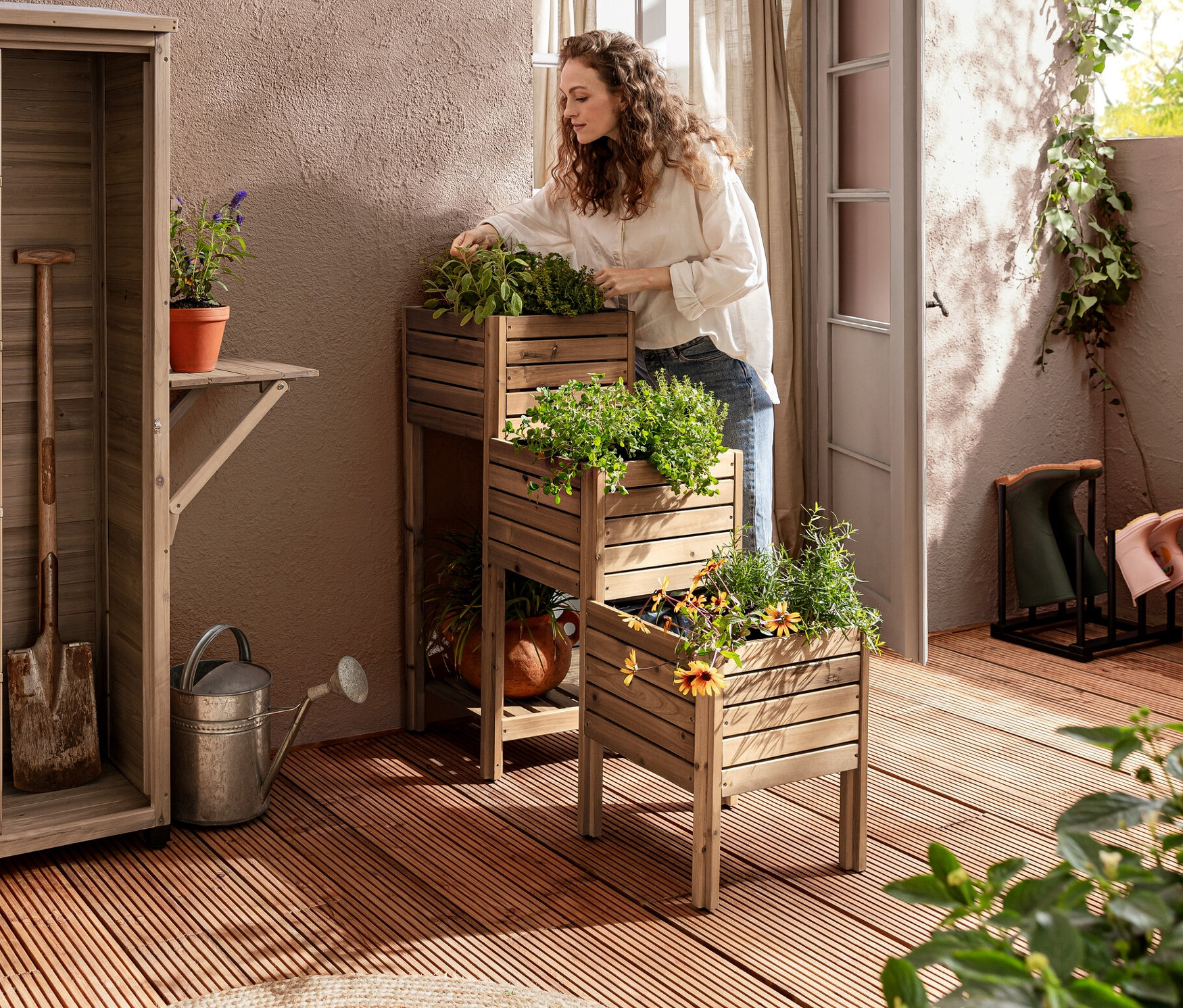 Auf einer Terrasse steht eine Frau an einer Pflanztreppe »Tinus« mit Regalboden, daneben ein großer Gartenschrank »Tinus« mit hochklappbarem Seitenregal.