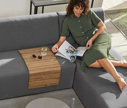 Eine Frau im grünen Kleid liest auf einer modularen Lounge-Ecke mit Teak-Sofatablett und 2 Premium-Outdoor-Kissen.