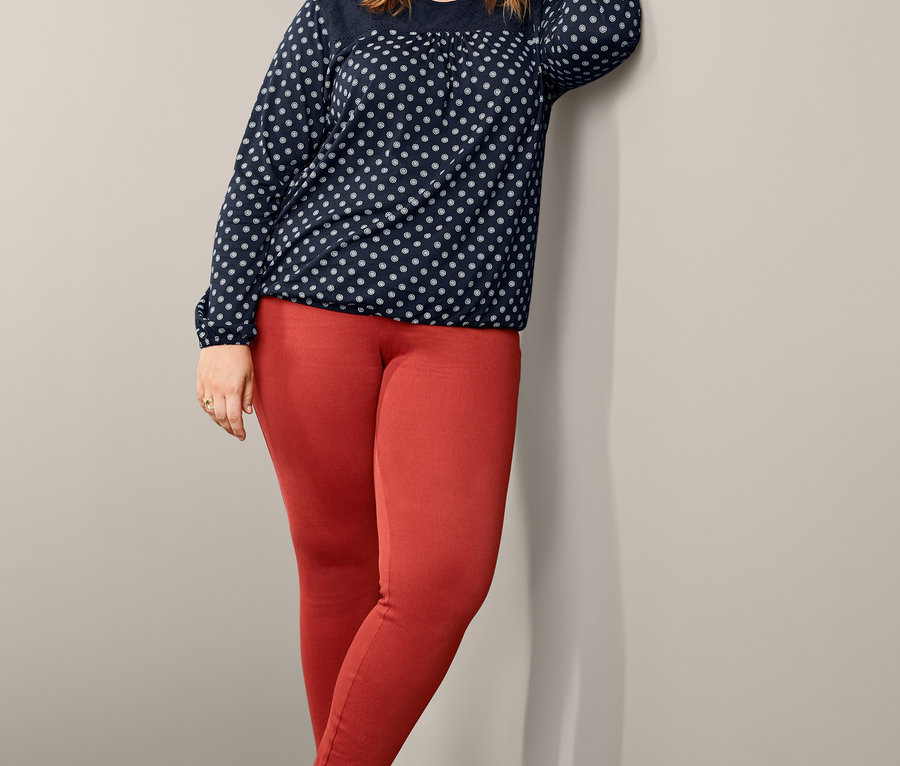 Eine Frau lehnt an einer Wand und trägt ein Blusenshirt mit Stickerei und Jeggings.