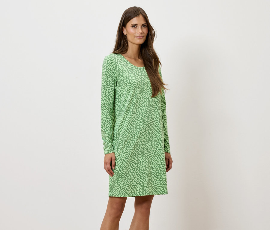 Eine Frau steht und posiert in einem grün gemusterten, langärmeligen Kleid.