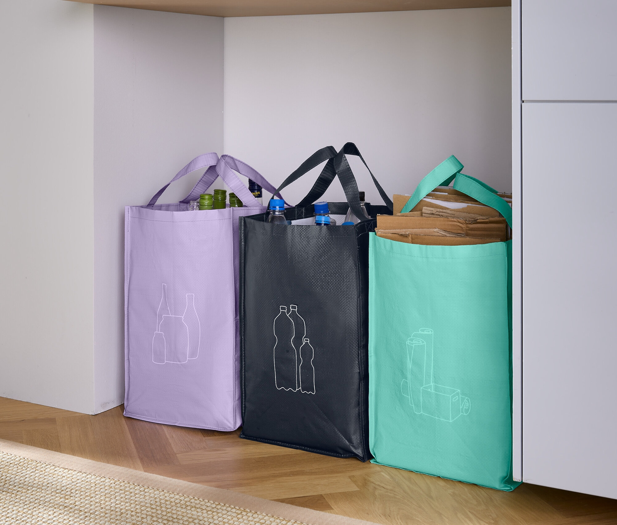 Drei Einkaufstaschen stehen nebeneinander: eine lila Tasche mit einer Zeichnung von Obst, eine schwarze Tasche mit einer Zeichnung von Flaschen und eine türkisblaue Tasche mit einer Zeichnung von einem Glas.