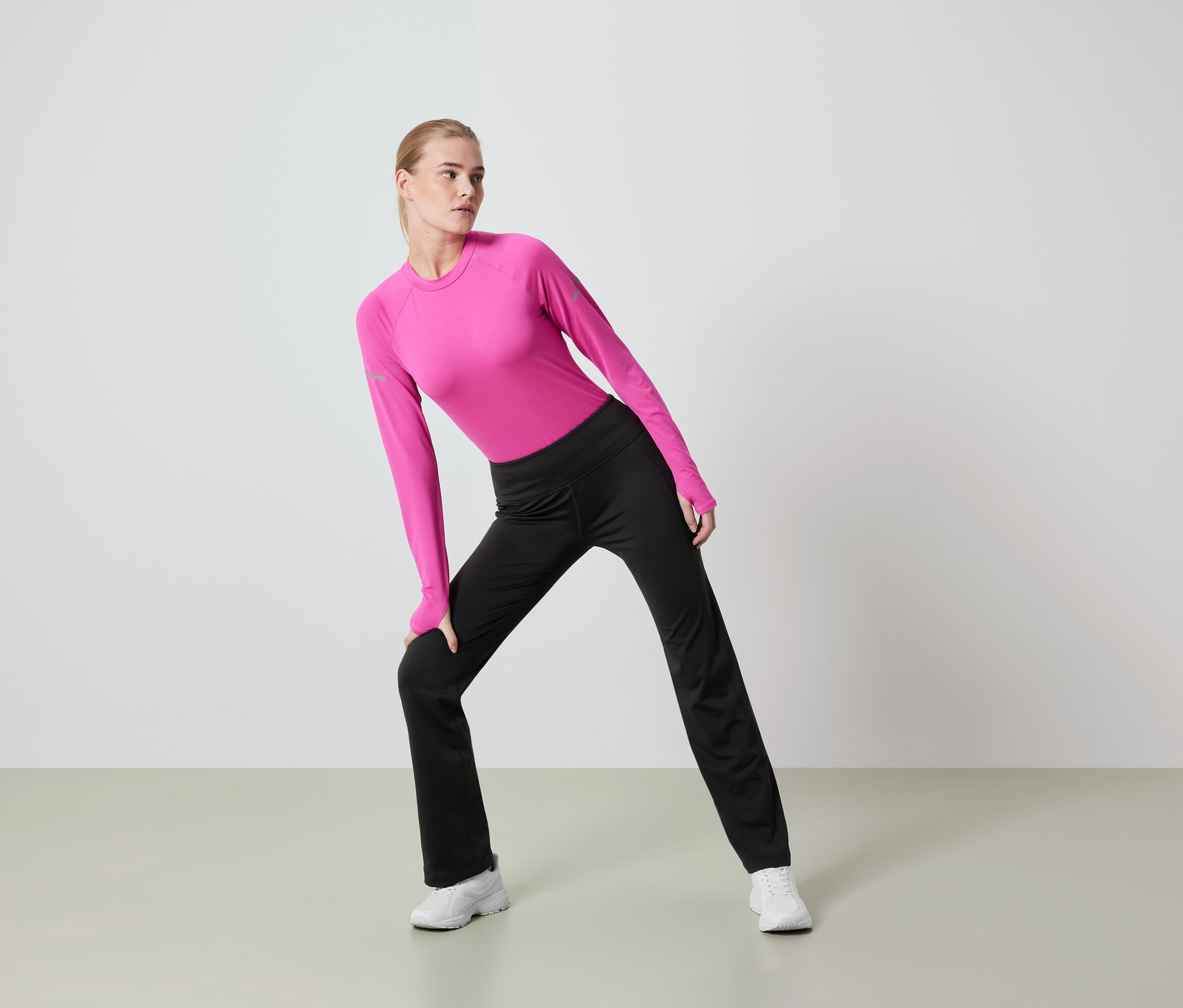 Frau mit blonden Haaren dehnt sich in einem pinkfarbenen Langarmshirt und einer schwarzen Hose.