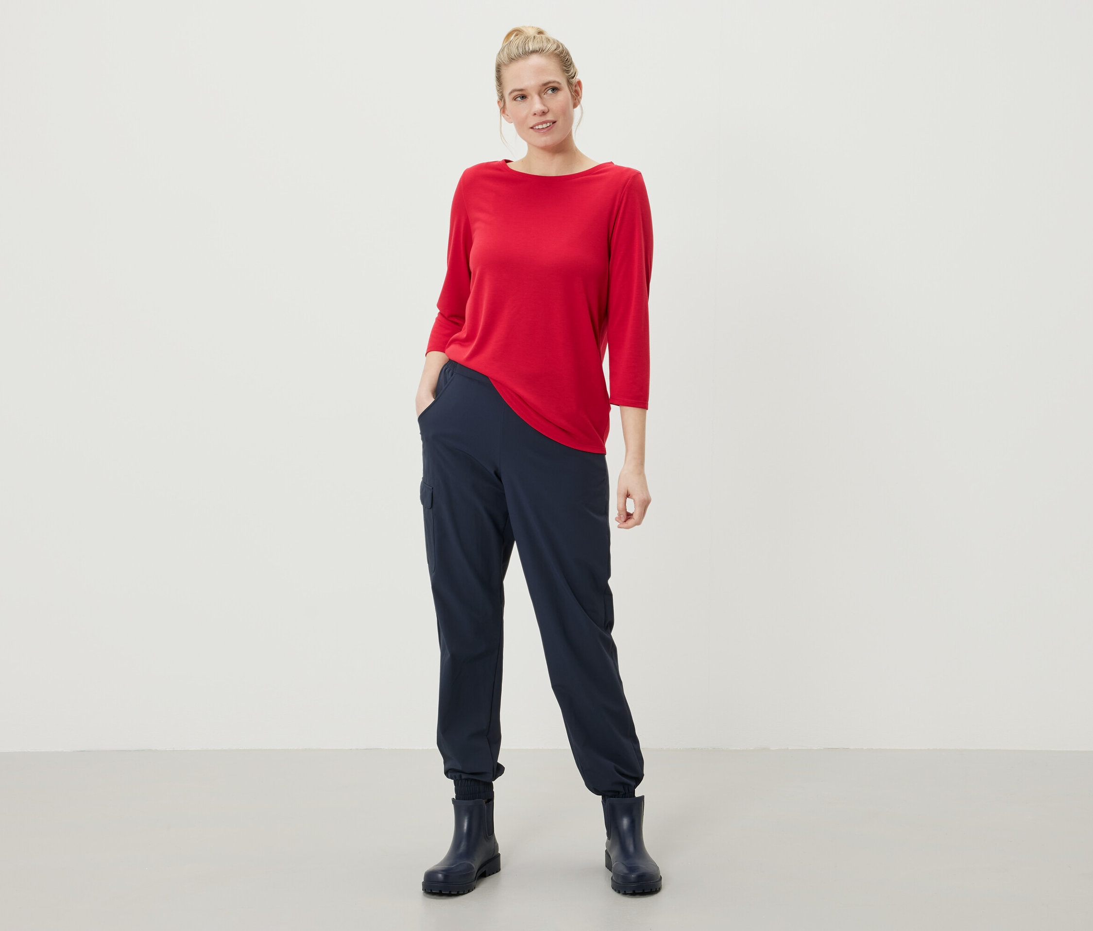 Eine Frau steht mit den Händen in den Taschen ihrer marineblauen Hose und trägt ein rotes Oberteil mit Dreiviertelärmeln und marineblaue Gummistiefel.