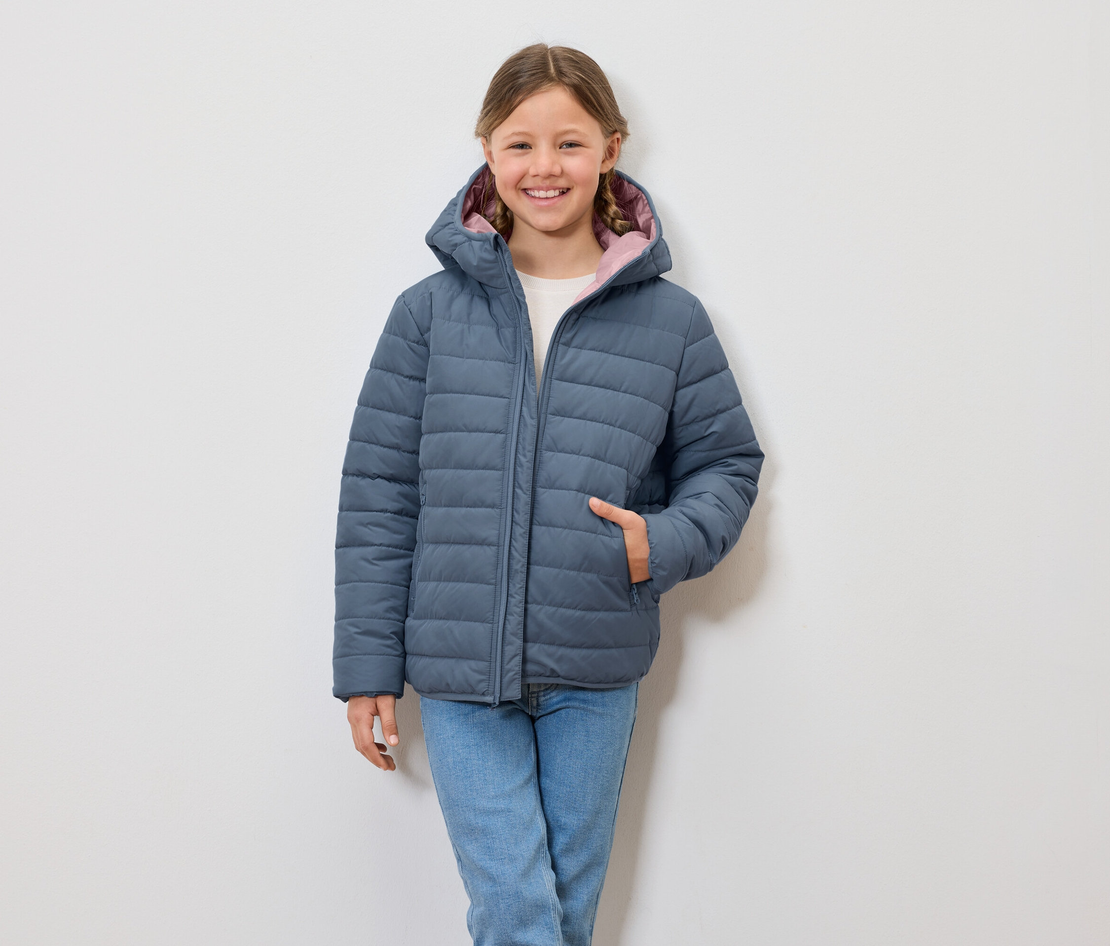 Lächelndes Mädchen trägt eine blaue Steppjacke mit Kapuze und Jeans.