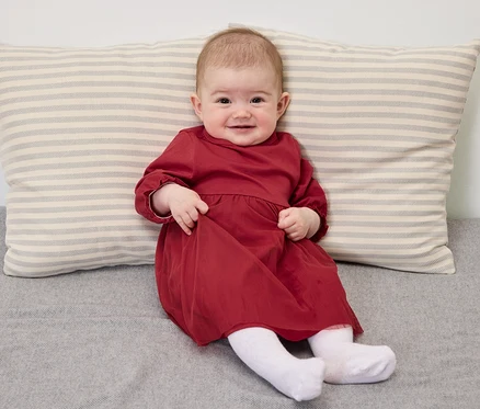 Ein lächelndes Baby in einem roten Kleid sitzt auf einer grauen Decke mit einem gestreiften Kissen.