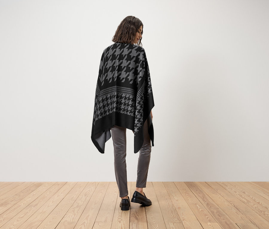 Frau trägt ein schwarz-graues Poncho mit Hahnentrittmuster, graue Hosen und schwarze Schuhe.