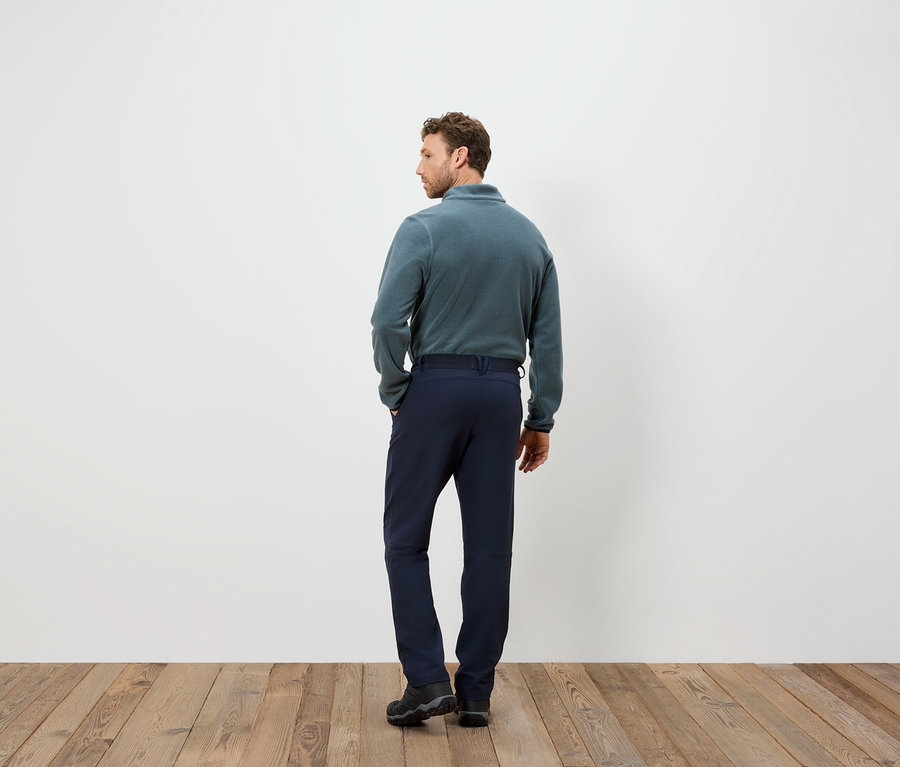 Mann in blauer Hose und Fleece-Pullover steht mit dem Rücken zur Kamera auf einem Holzboden.