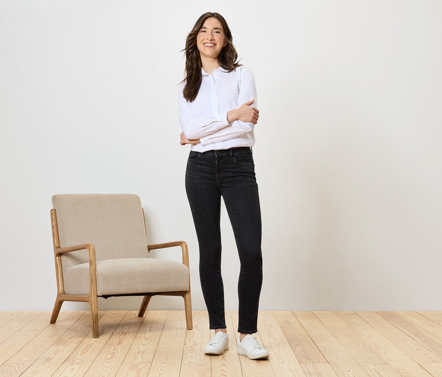 Eine Frau steht mit verschränkten Armen neben einem beigen Sessel und trägt schwarze Jeans und ein weißes Hemd.