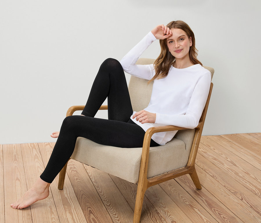 Eine Frau sitzt in einem Sessel und trägt ein weißes Langarmshirt und schwarze Leggings.