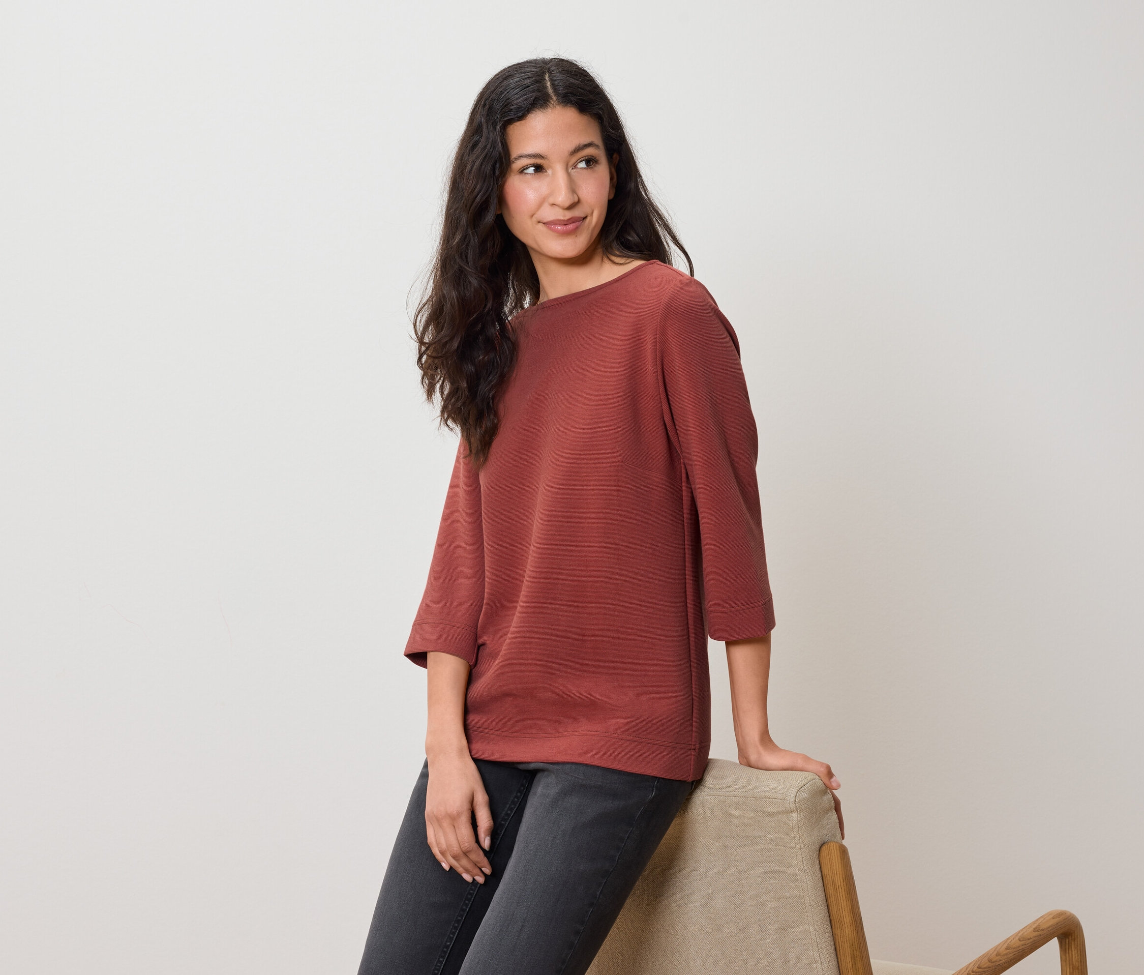 Eine Frau sitzt auf einem Stuhl und trägt ein rotbraunes Oberteil mit Dreiviertelärmeln und dunkle Jeans.