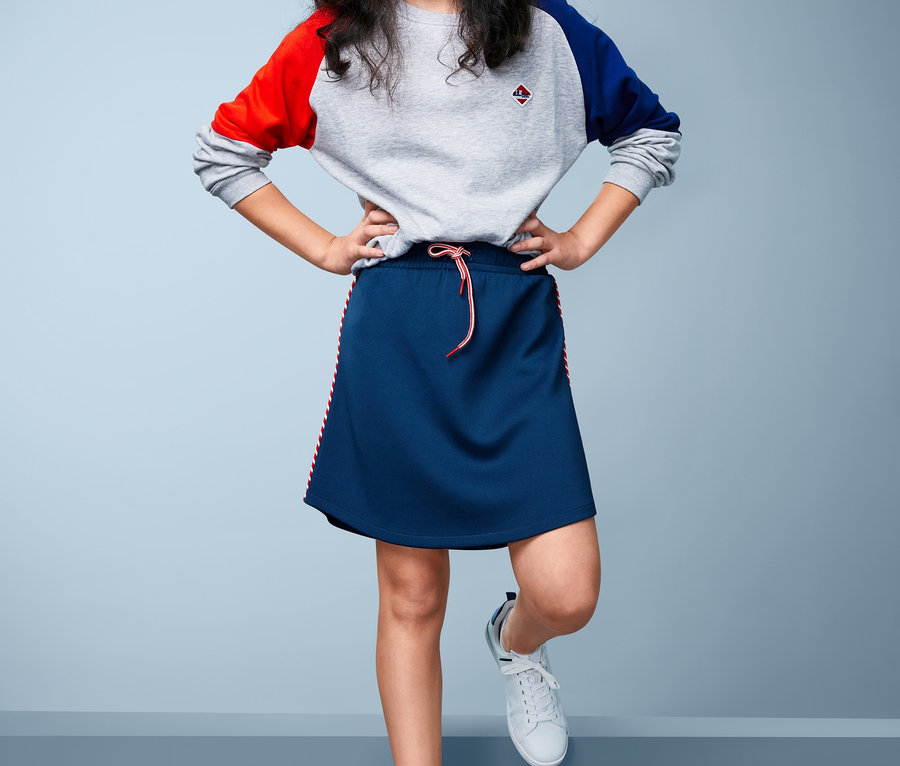 Mädchen steht mit den Händen in den Hüften und trägt einen sportlichen Jerseyrock und einen Pullover mit farbigen Ärmeln.