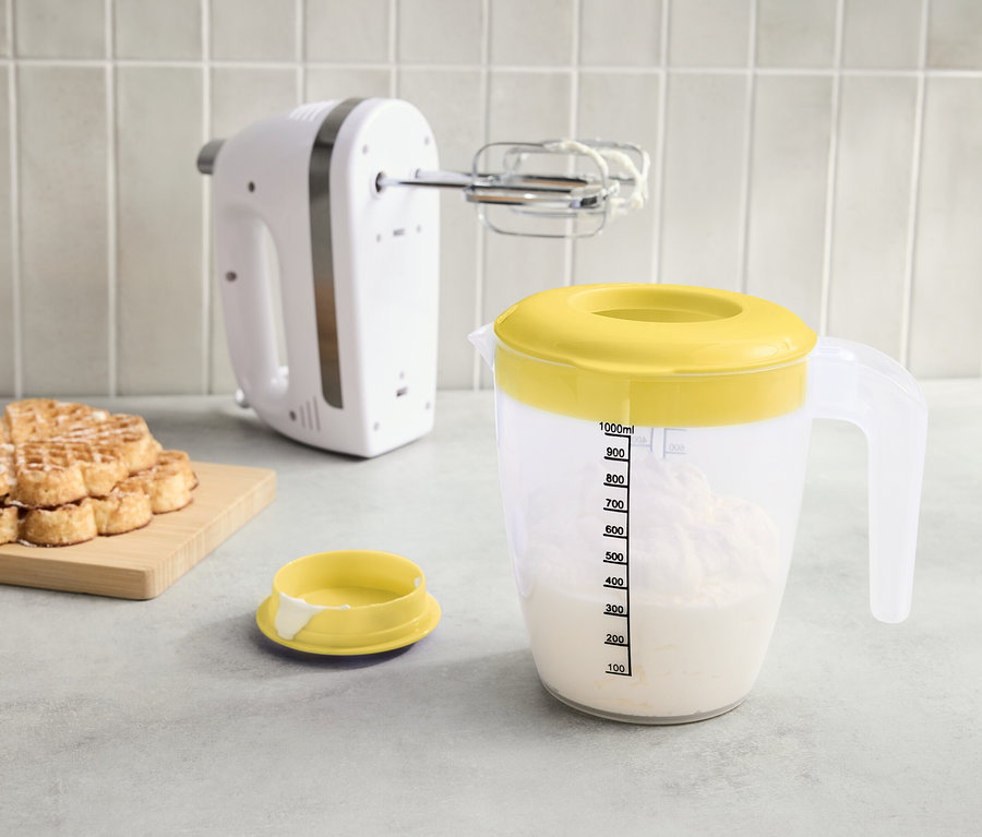 Ein weißer Mixer, ein Messbecher mit gelbem Deckel voller Flüssigkeit und ein Stapel Waffeln auf einem Brett stehen auf der Arbeitsfläche.