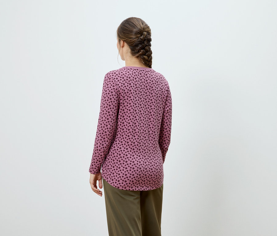 Rückansicht einer Frau mit braunem Zopf, die ein rosafarbenes Langarmshirt mit dunklem Muster und Khakihose trägt.