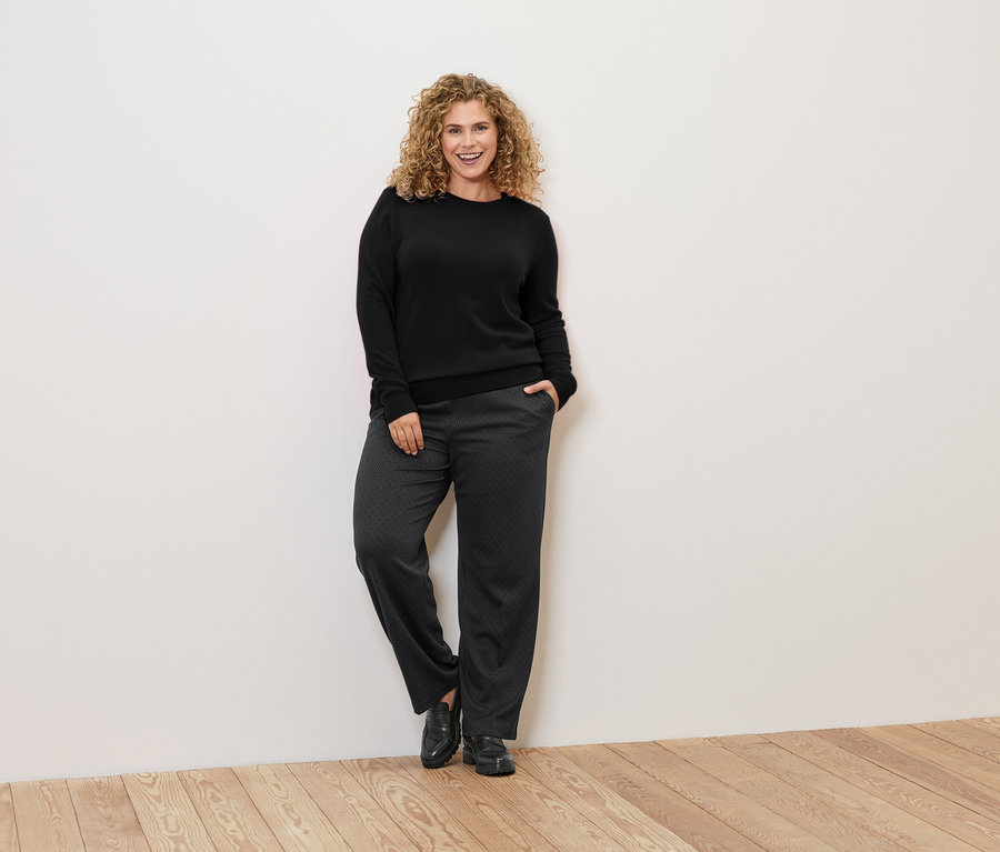 Eine Frau mit lockigem Haar lehnt an einer Wand und trägt einen schwarzen Pullover und eine gemusterte Hose.