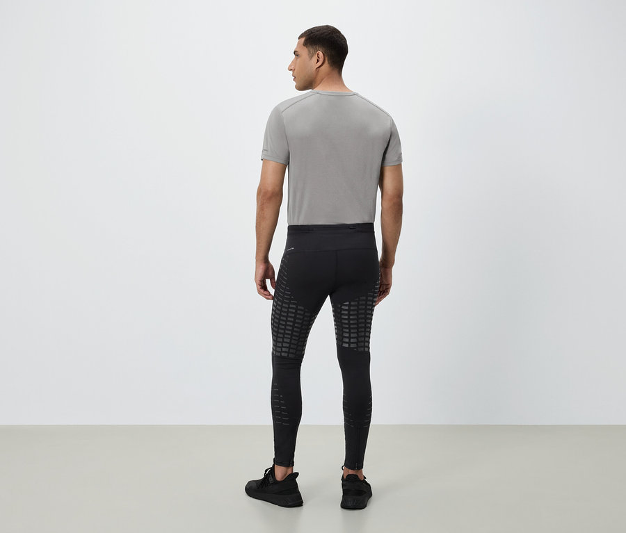 Mann von hinten trägt ein graues T-Shirt, eine schwarze Leggings mit reflektierenden Quadraten und schwarze Turnschuhe.