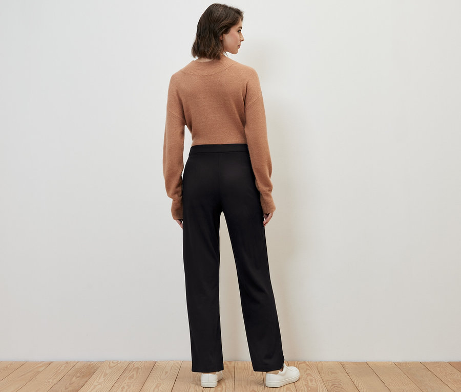 Eine Frau steht mit dem Rücken zur Kamera und trägt eine Jerseyhose mit Biese, einen braunen Pullover und weiße Turnschuhe.