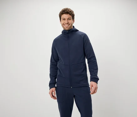 Ein lächelnder Mann posiert in einem blauen Trainingsanzug mit Kapuzenjacke und Hose vor einem weißen Hintergrund.