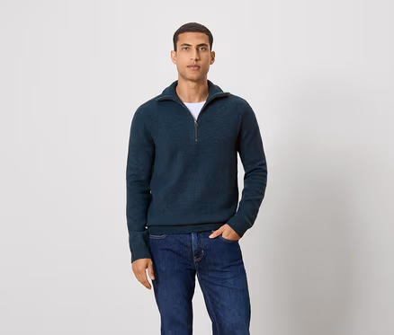 Mann posiert vor einem Hintergrund und trägt einen dunkelblauen Pullover mit Reißverschluss und Jeans.