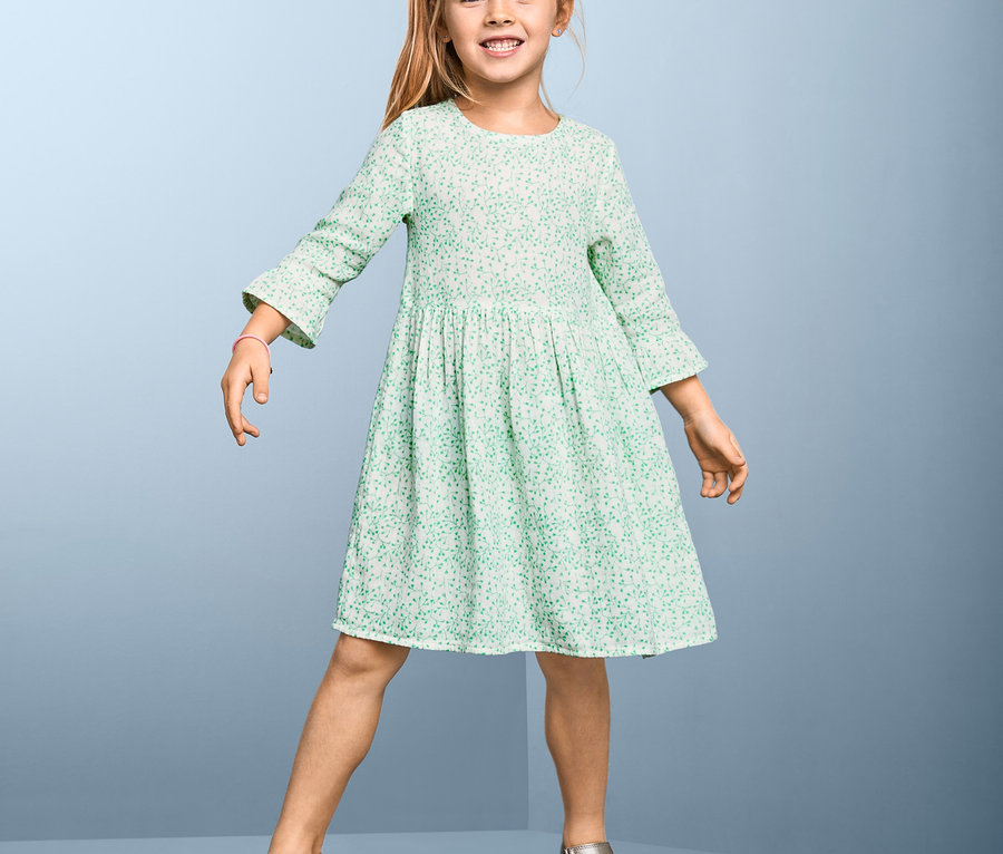Lächelndes Mädchen posiert in einem Baumwoll-Webkleid mit Blumenmuster und Sandalen.