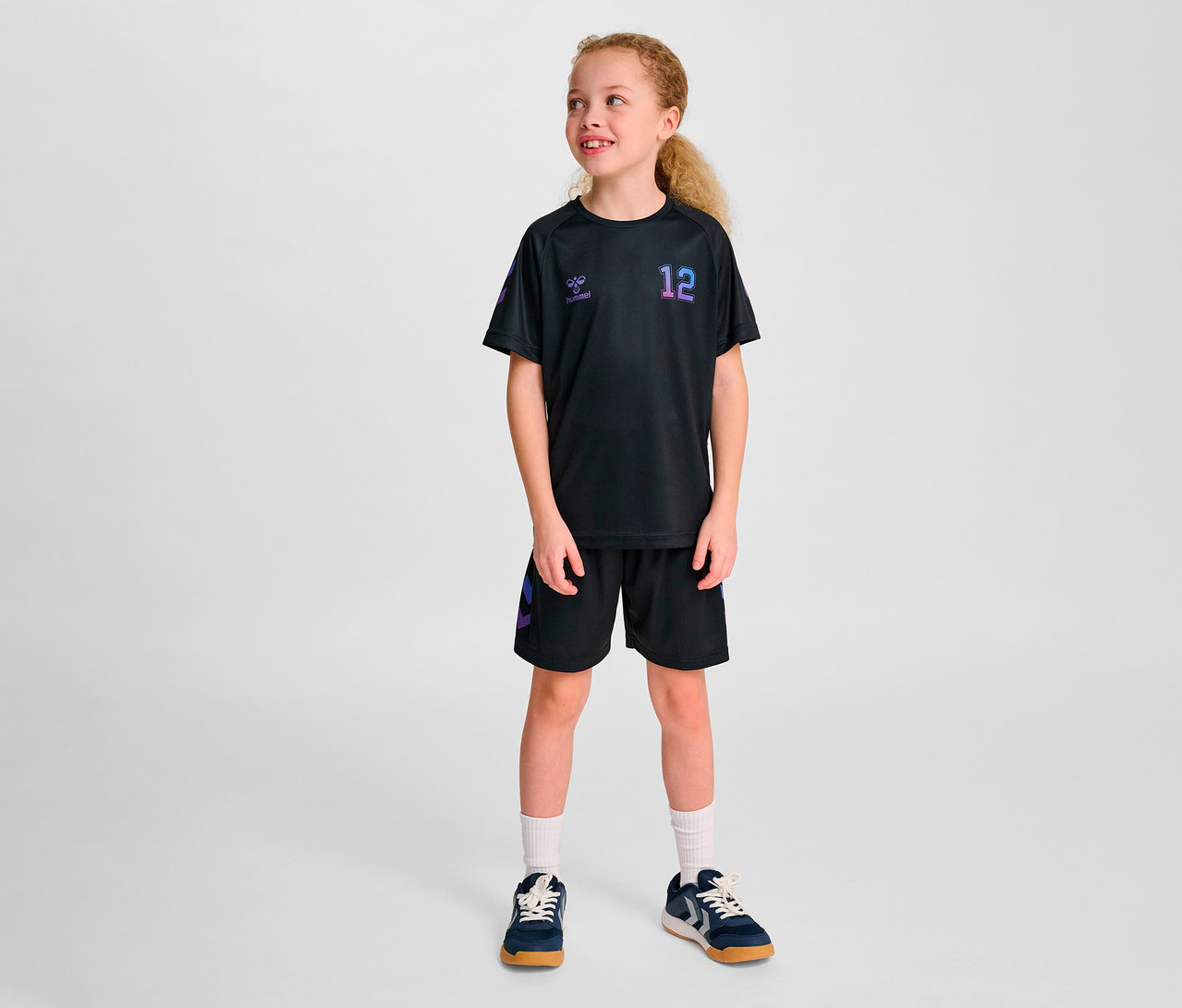 Ein Mädchen posiert in schwarzer Sportbekleidung, bestehend aus einem Hummel HMLJR shimmer T-Shirt S/S und Shorts.